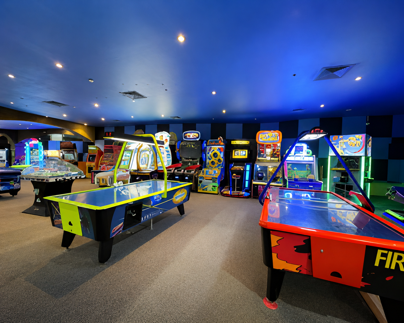 Indoor arcade floor with two air hockey tables, a glass-front prize redemption counter and a row of upright arcade and ticket-redemption cabinets under a blue-lit ceiling with a checkerboard accent wall