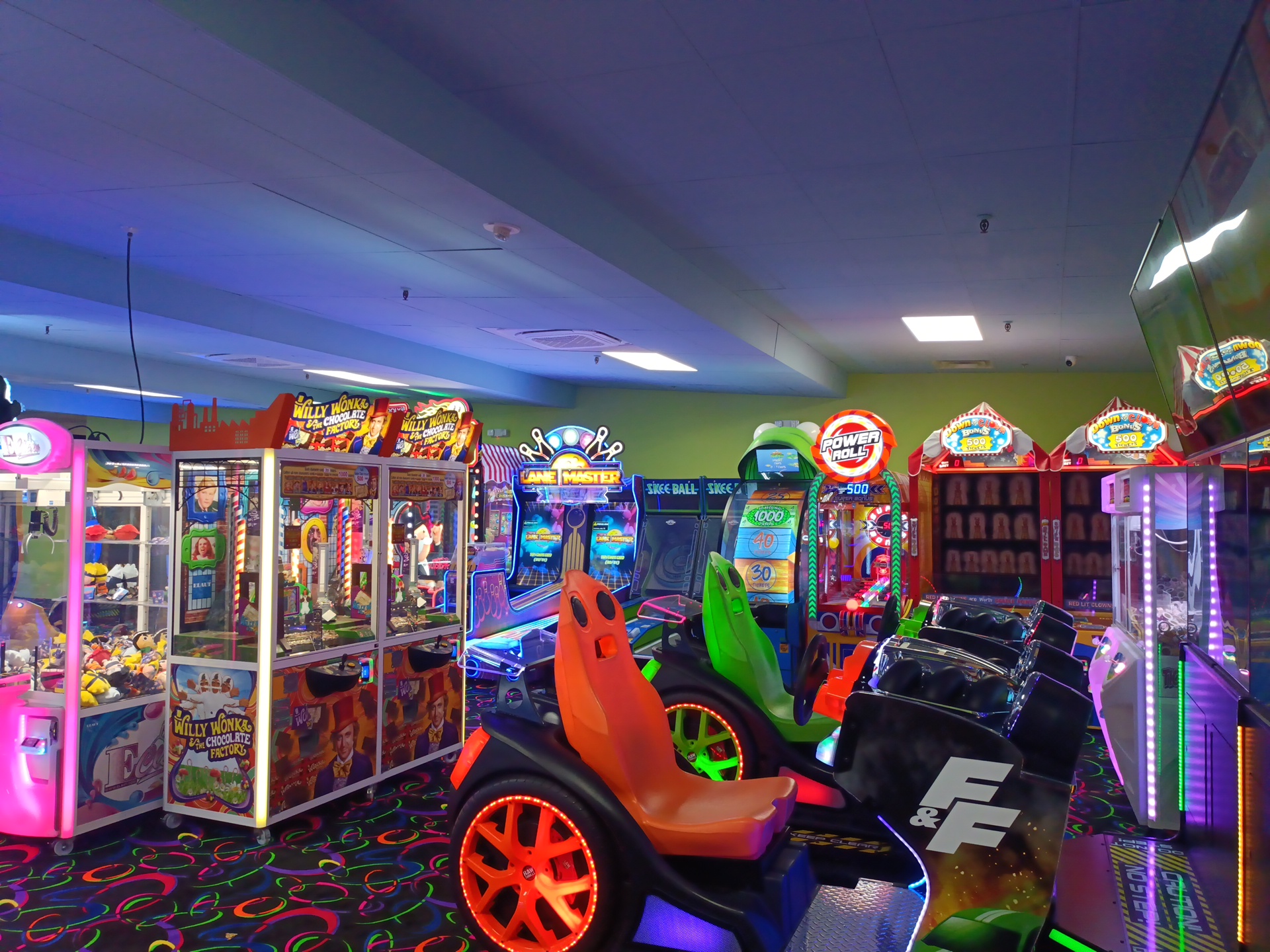 US family arcade game room with plush crane machines, ticket-redemption cabinets, skill-roller alleys and twin kids' racing simulators on patterned carpet