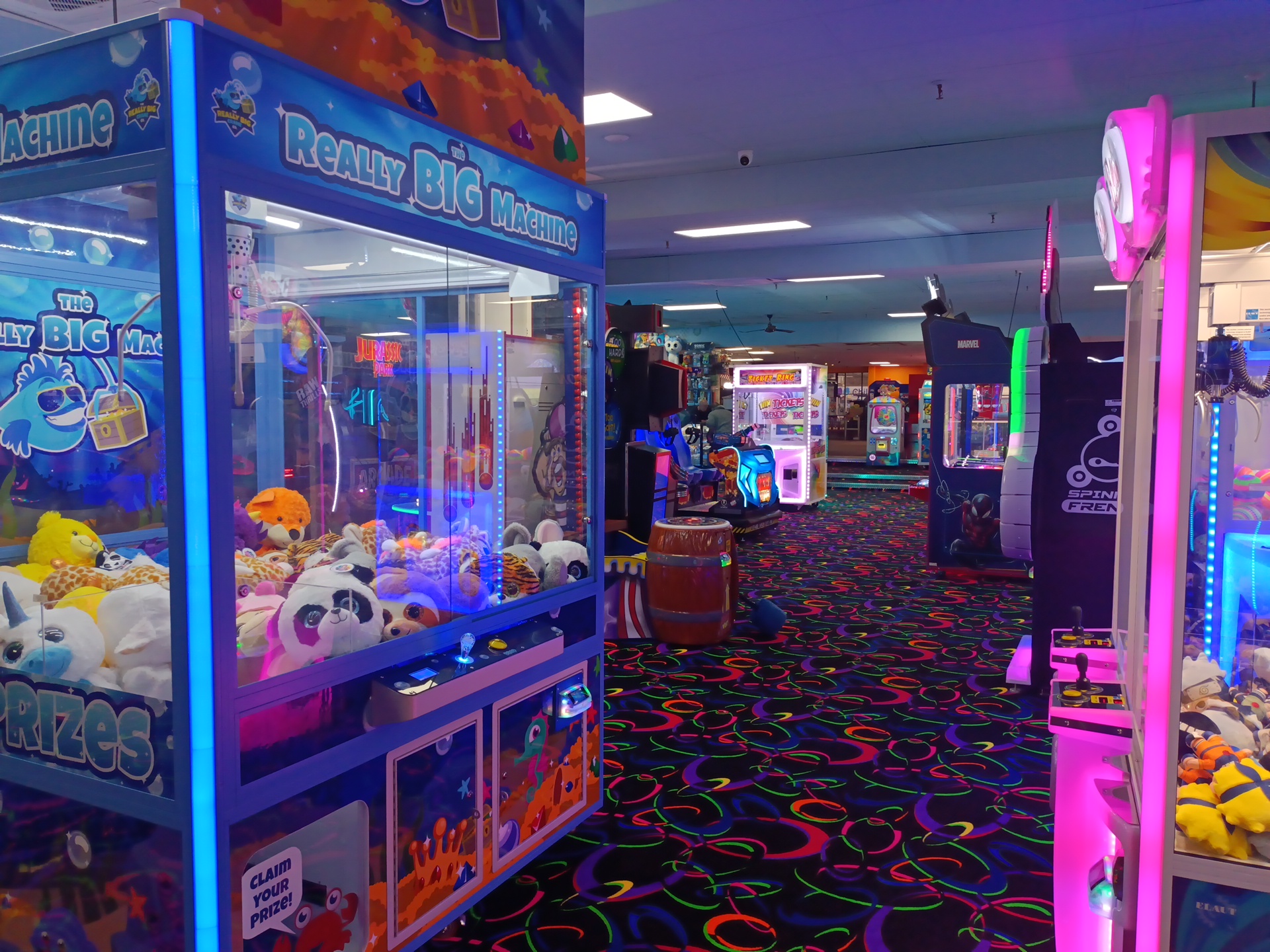US indoor arcade prize zone: a large plush claw crane machine full of soft toys, pink-neon claw cabinets, ticket-redemption games, kids' racing simulators and a barrel ride on neon-swirl carpet.