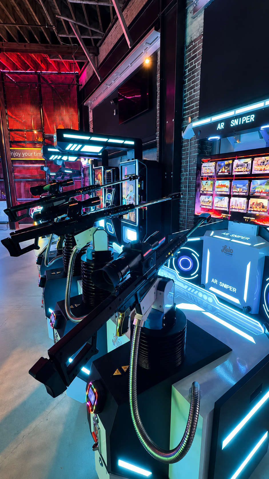Row of AR Sniper shooting simulator arcade cabinets with large rifle controllers and game-select LCD screens in an indoor family entertainment centre in Saudi Arabia, with a red climbing-net play structure behind