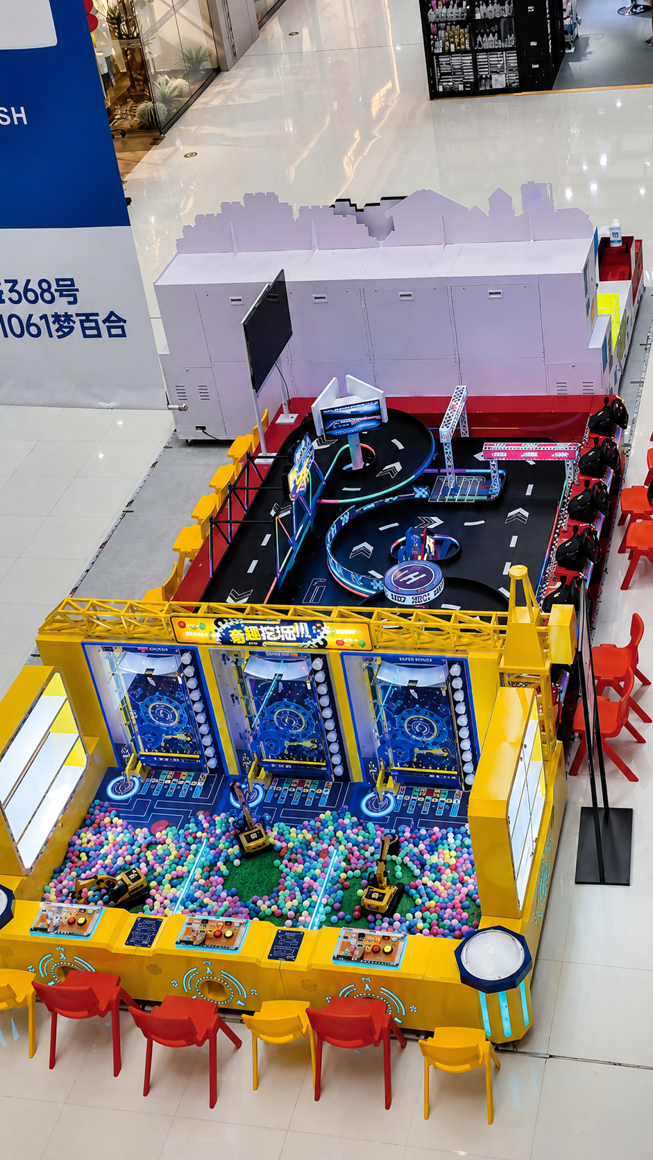 Overhead view of a compact children's play zone in a Chinese shopping mall: three ball-pit excavator claw-digger stations over a pit of colorful plastic balls, next to a neon remote-control car racing track with checkered start arch and red and yellow kids' chairs